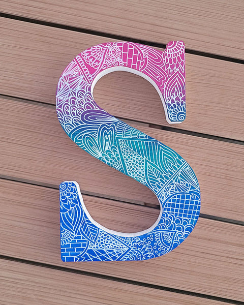 A gradient hand painted letter S (pink-green-blue) with doodles in white