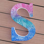 A gradient hand painted letter S (pink-green-blue) with doodles in white