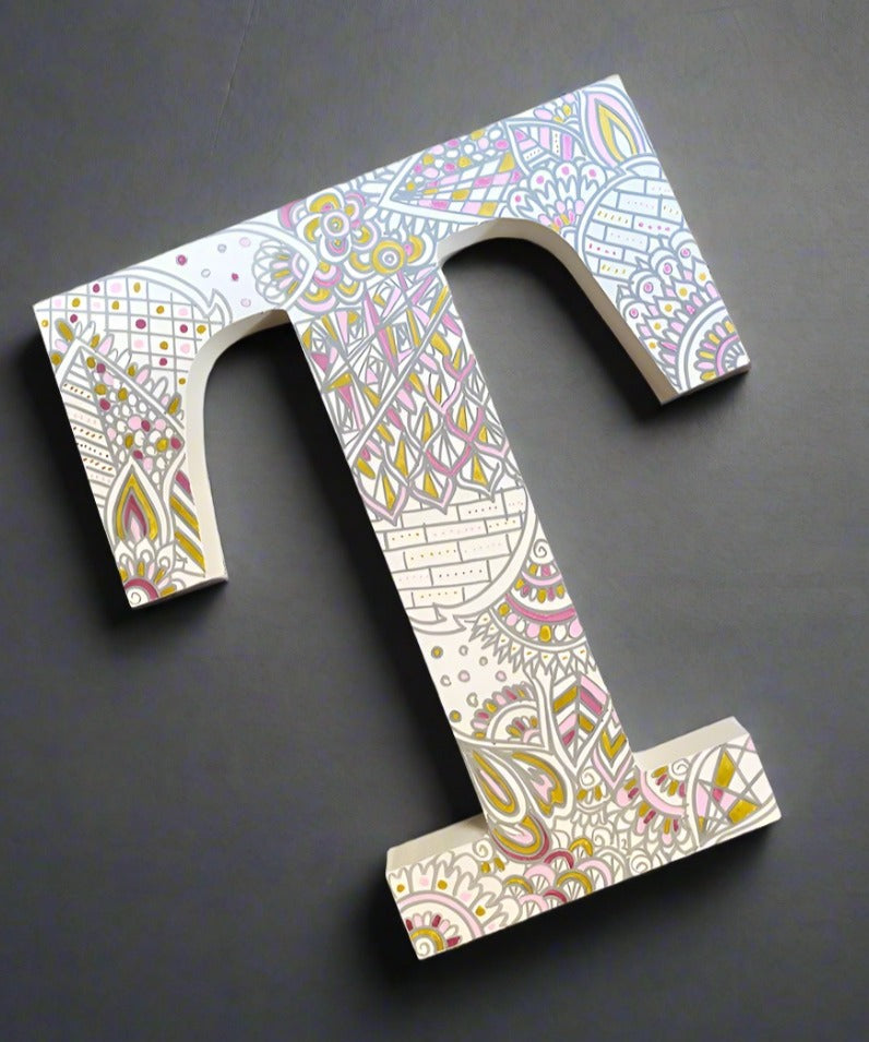 A White letter T with doodles drawn in grey and colored in with gold, shiny pink and baby pink