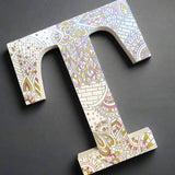 A White letter T with doodles drawn in grey and colored in with gold, shiny pink and baby pink