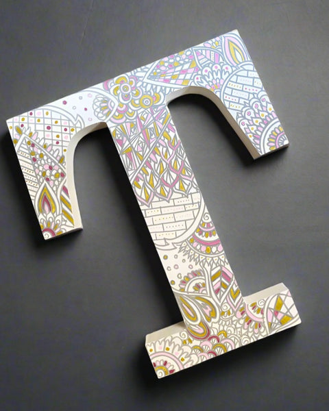 A White letter T with doodles drawn in grey and colored in with gold, shiny pink and baby pink