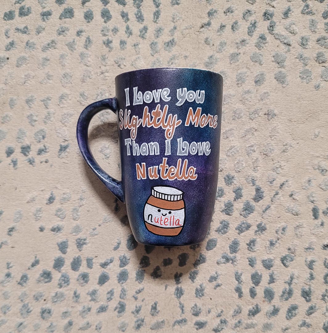 A black sky hand painted mug with the sentence "I love you slightly more than I love Nutella"and a small Nutella jar Painted with a smiley face