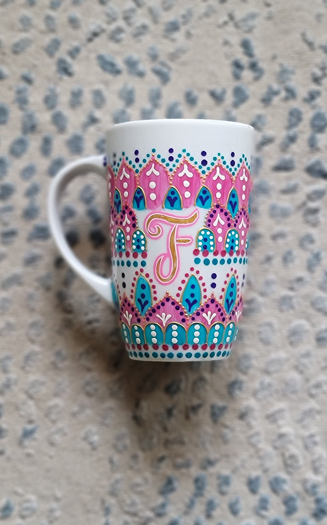 A White mug with patterns in pink and turquoise outlined with gold relief and dot art of white purple pink and turquoise, with the letter F written in the middle