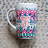 A White mug with patterns in pink and turquoise outlined with gold relief and dot art of white purple pink and turquoise, with the letter F written in the middle