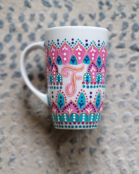 A White mug with patterns in pink and turquoise outlined with gold relief and dot art of white purple pink and turquoise, with the letter F written in the middle