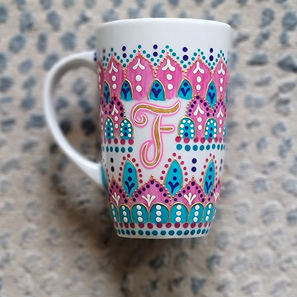 A White mug with patterns in pink and turquoise outlined with gold relief and dot art of white purple pink and turquoise, with the letter F written in the middle