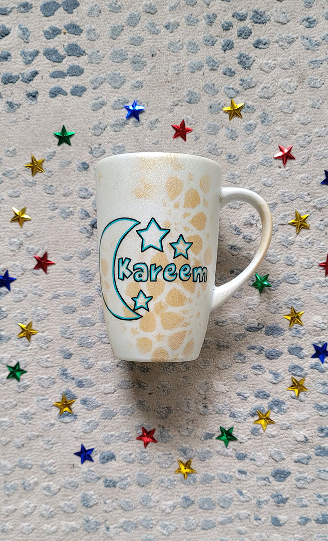 A Shimmery White with gold islamic patterns hand painted mug with a moon painted in black and blue green and the name Kareem beside it