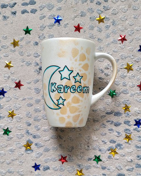 A Shimmery White with gold islamic patterns hand painted mug with a moon painted in black and blue green and the name Kareem beside it