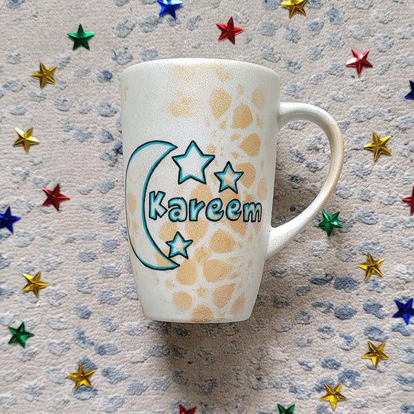 A Shimmery White with gold islamic patterns hand painted mug with a moon painted in black and blue green and the name Kareem beside it