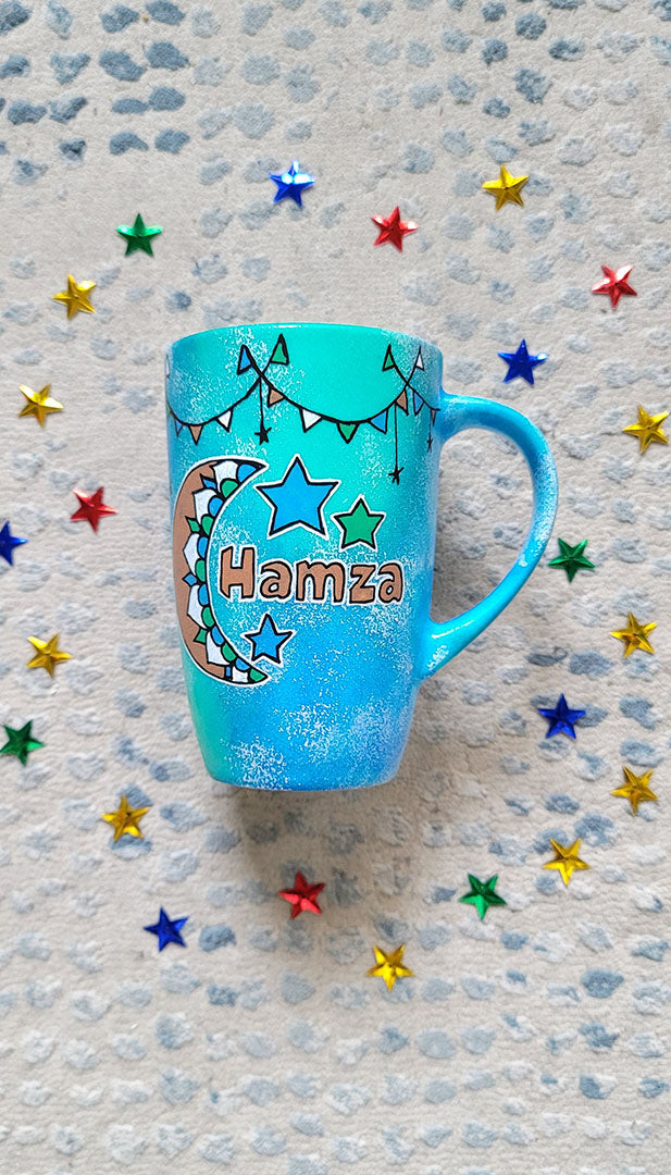 A Blue Green Hand painted mug with decorations hanging from the top all around the mug and a moon painted in black brown, blue and green and the name Hamza written beside it