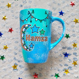 A Blue Green Hand painted mug with decorations hanging from the top all around the mug and a moon painted in black brown, blue and green and the name Hamza written beside it