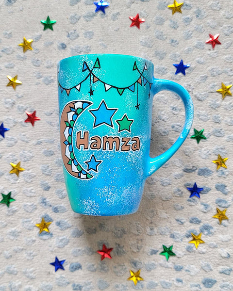 A Blue Green Hand painted mug with decorations hanging from the top all around the mug and a moon painted in black brown, blue and green and the name Hamza written beside it