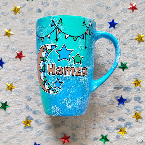 A Blue Green Hand painted mug with decorations hanging from the top all around the mug and a moon painted in black brown, blue and green and the name Hamza written beside it