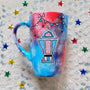 A Blue Red Hand painted mug with decorations hanging from the top all around the mug and a fanoos painted in black grey, blue and red