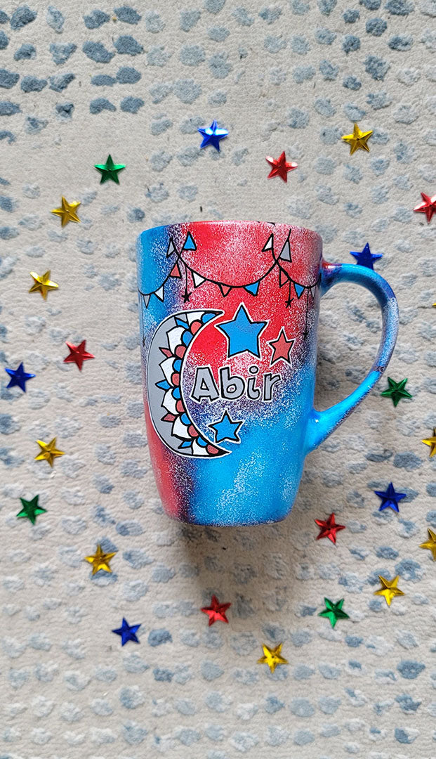 A Blue Red Hand painted mug with decorations hanging from the top all around the mug and a moon painted in black grey, blue and red and the name abir beside it