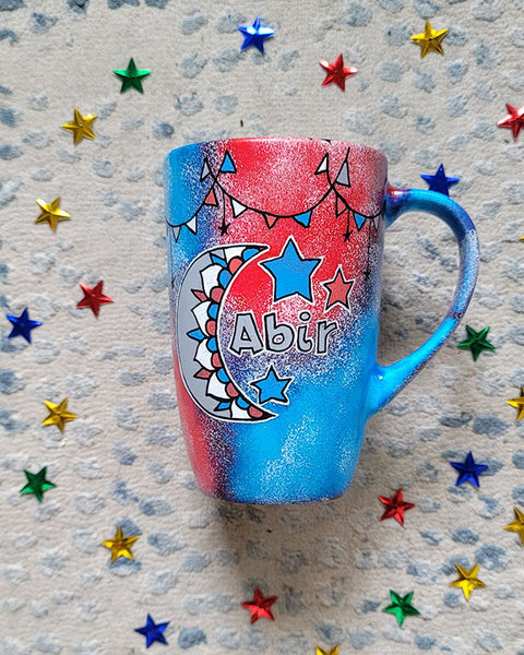 A Blue Red Hand painted mug with decorations hanging from the top all around the mug and a moon painted in black grey, blue and red and the name abir beside it