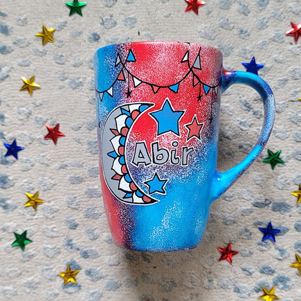 A Blue Red Hand painted mug with decorations hanging from the top all around the mug and a moon painted in black grey, blue and red and the name abir beside it