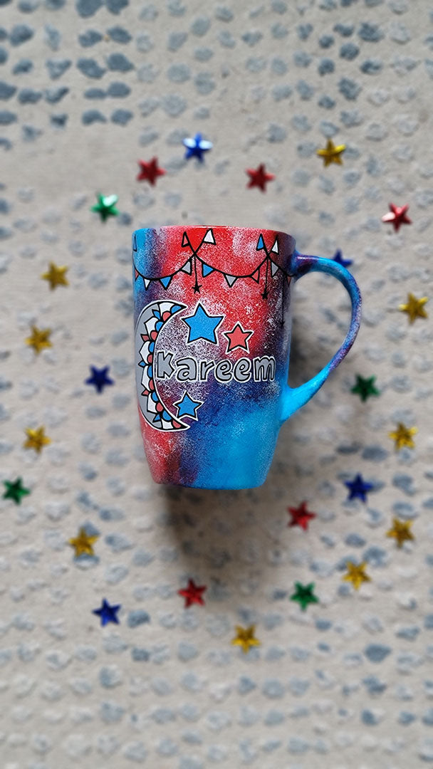 A Blue Red Hand painted mug with decorations hanging from the top all around the mug and a moon painted in black grey, blue and red and the name Kareem beside it