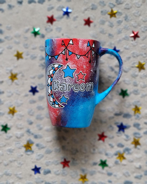 A Blue Red Hand painted mug with decorations hanging from the top all around the mug and a moon painted in black grey, blue and red and the name Kareem beside it