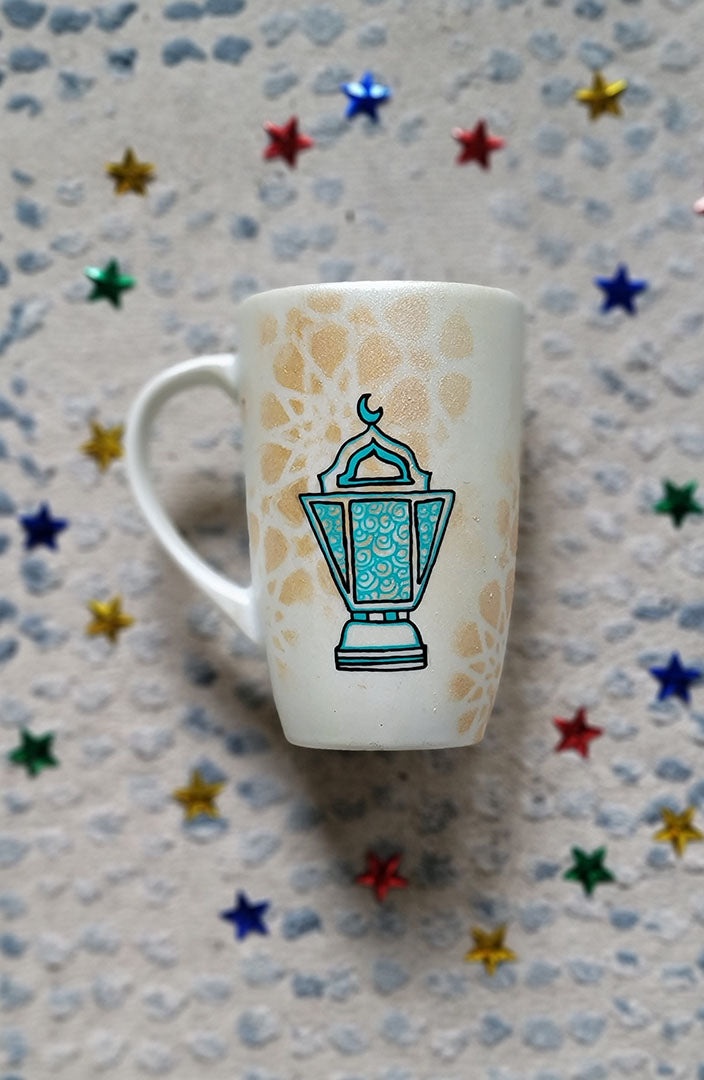 A Shimmery White with gold islamic patterns hand painted mug with a fanoos painted in black and blue green patterns