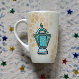 A Shimmery White with gold islamic patterns hand painted mug with a fanoos painted in black and blue green patterns