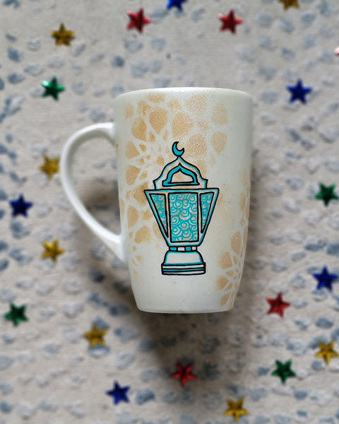 A Shimmery White with gold islamic patterns hand painted mug with a fanoos painted in black and blue green patterns