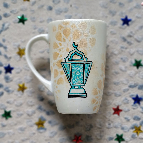 A Shimmery White with gold islamic patterns hand painted mug with a fanoos painted in black and blue green patterns