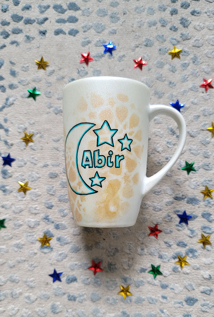 A Shimmery White with gold islamic patterns hand painted mug with a moon painted in black and blue green and the name Abir beside it