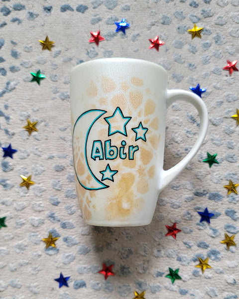 A Shimmery White with gold islamic patterns hand painted mug with a moon painted in black and blue green and the name Abir beside it