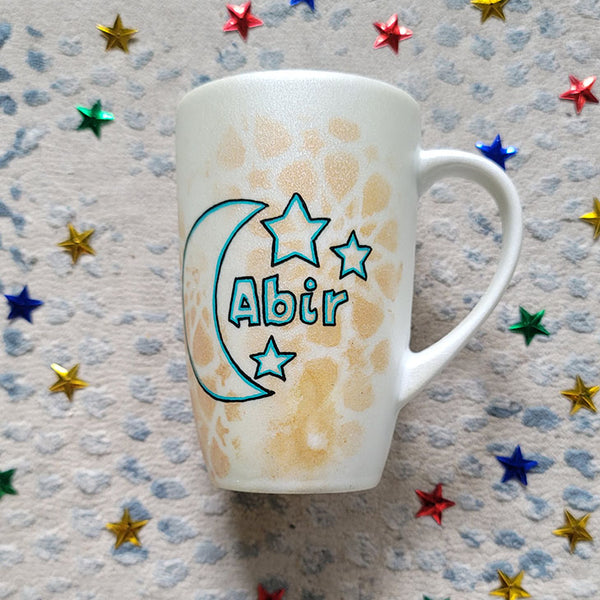 A Shimmery White with gold islamic patterns hand painted mug with a moon painted in black and blue green and the name Abir beside it