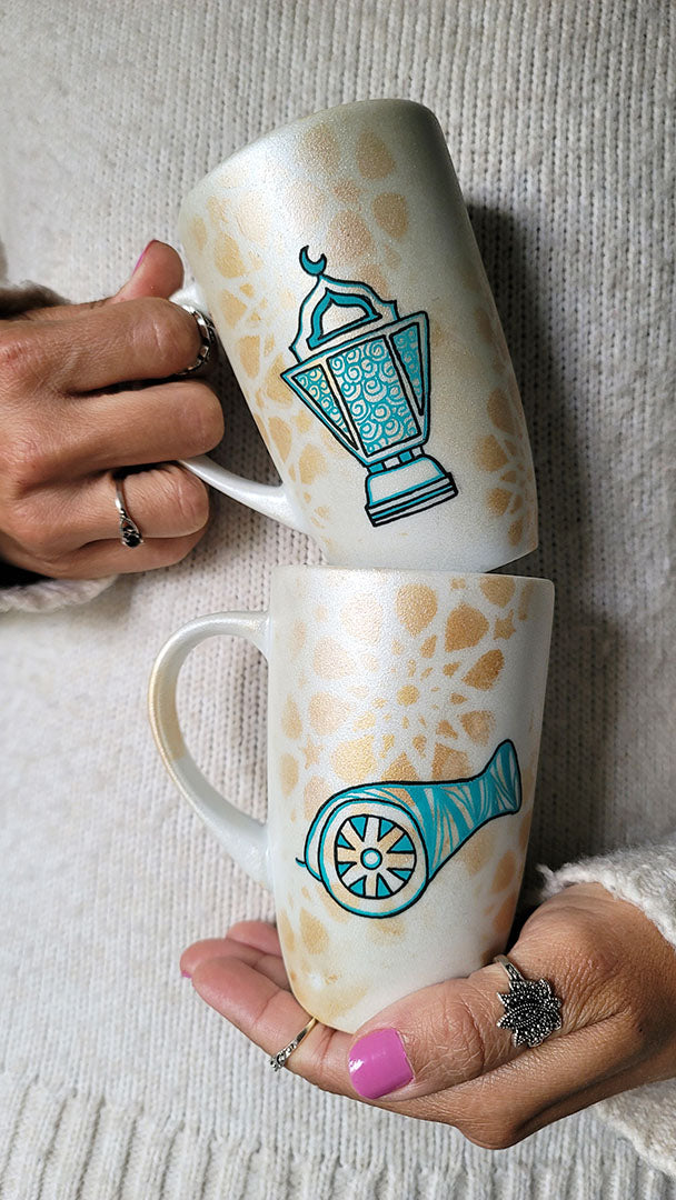 2 hand painted mugs in white shimmery color and faded gold islamic patterns one with a fanoos and the other with a madfa3