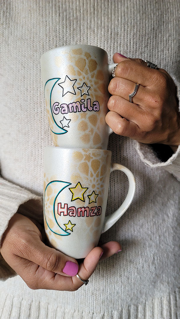 2 shimmery white hand painted mugs with a faded geometric pattern in gold, one with the name gamila and a moon and the other with the name hamza and a moon