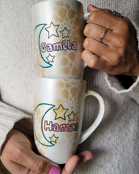 2 shimmery white hand painted mugs with a faded geometric pattern in gold, one with the name gamila and a moon and the other with the name hamza and a moon