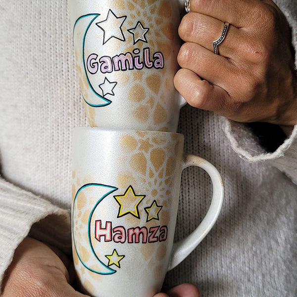 2 shimmery white hand painted mugs with a faded geometric pattern in gold, one with the name gamila and a moon and the other with the name hamza and a moon