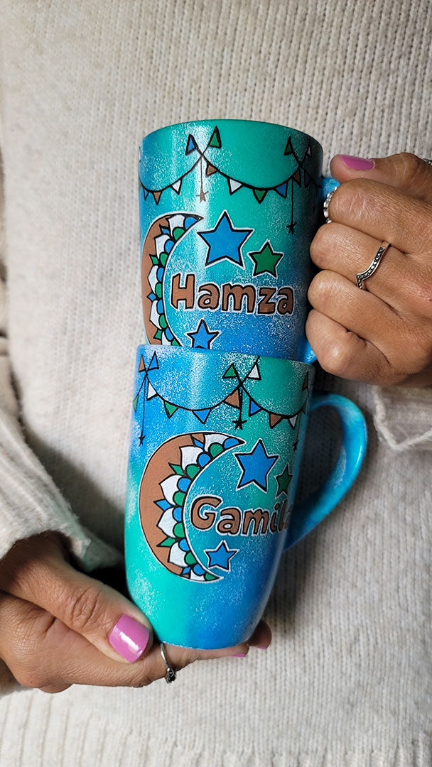 2 hand painted mugs in blue green color with decorations all around the top and one has a a moon with the name hamza and the other a moon with the name gamila