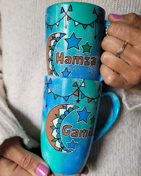 2 hand painted mugs in blue green color with decorations all around the top and one has a a moon with the name hamza and the other a moon with the name gamila