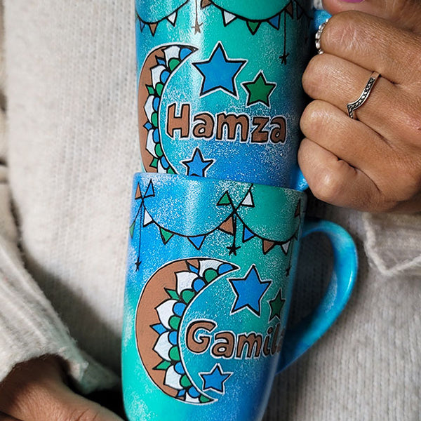 2 hand painted mugs in blue green color with decorations all around the top and one has a a moon with the name hamza and the other a moon with the name gamila