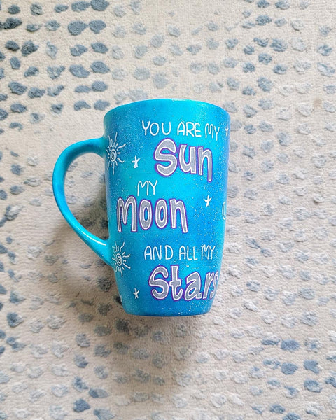 A Blue Galaxy Hand painted mug with the sentence "you are my Sun, My Moon and all my stars" with small moons stars and suns scattered all over the mug