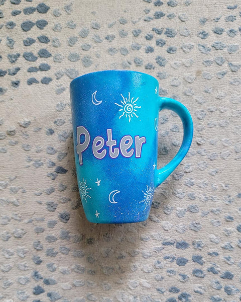 A Blue Galaxy hand painted mug with the name "Peter" with small moons stars and suns scattered all over.