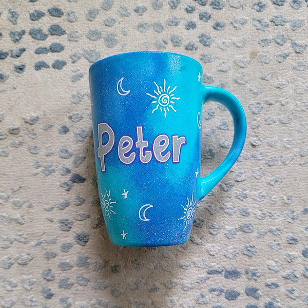 A Blue Galaxy hand painted mug with the name "Peter" with small moons stars and suns scattered all over.