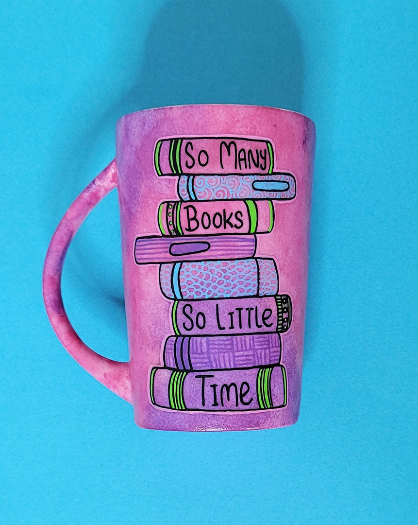 A Pink hand painted mug with books on top of each other and words written in between making the sentence" So Many books, So little time"