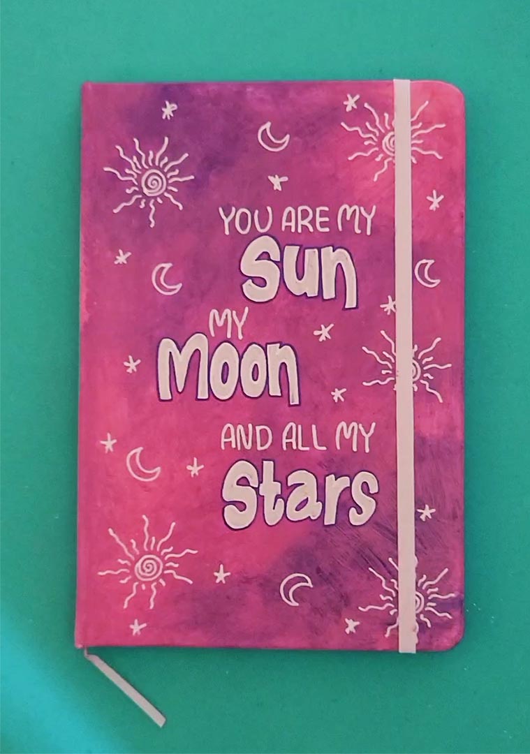 A Pink Galaxy Hand painted Notebook with the sentence "you are my Sun, My Moon and all my stars" with small moons stars and suns scattered all over the mug