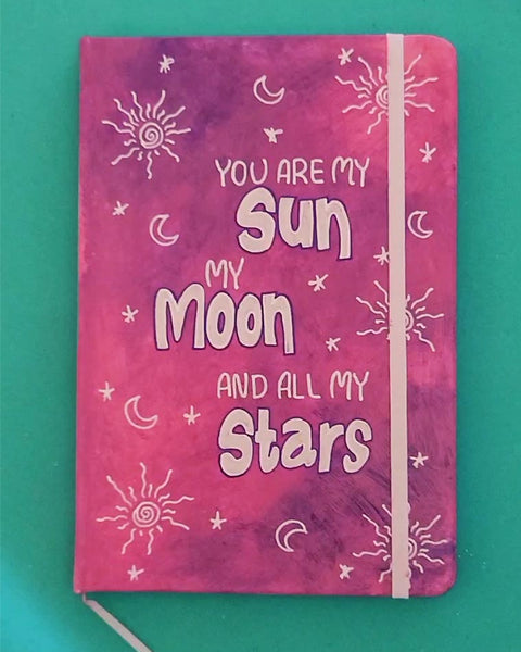 A Pink Galaxy Hand painted Notebook with the sentence "you are my Sun, My Moon and all my stars" with small moons stars and suns scattered all over the mug