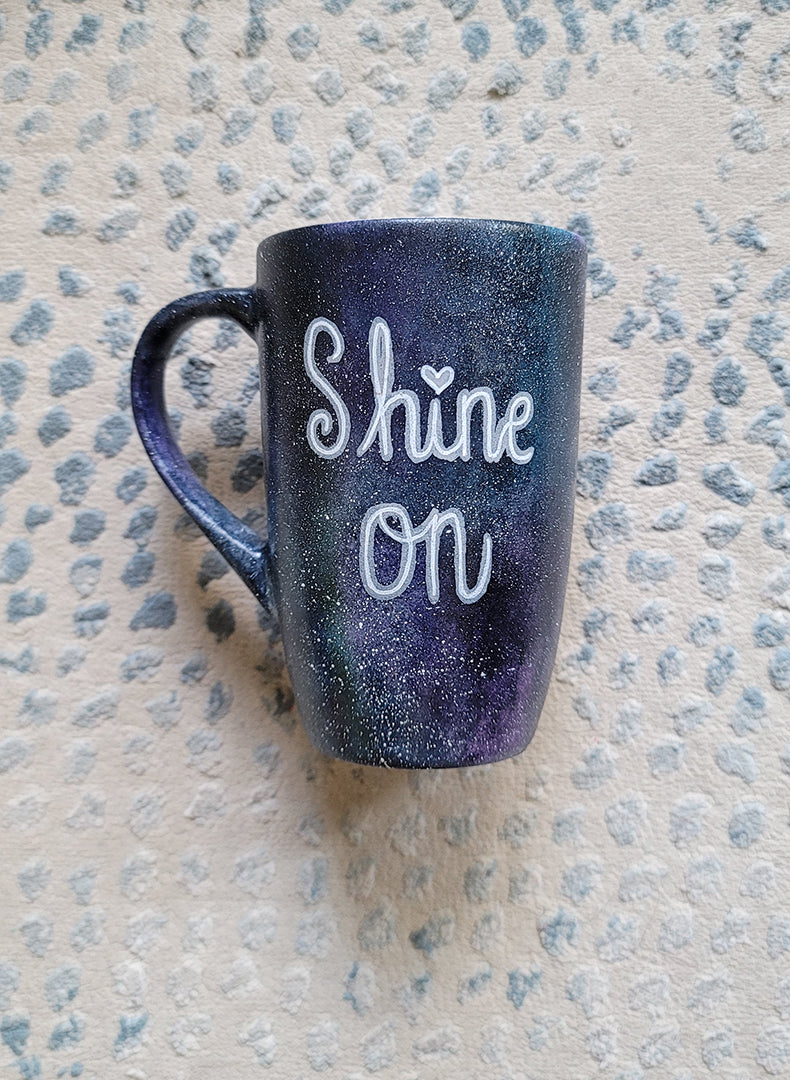 A Black galaxy hand painted mug with the words "Shine on"