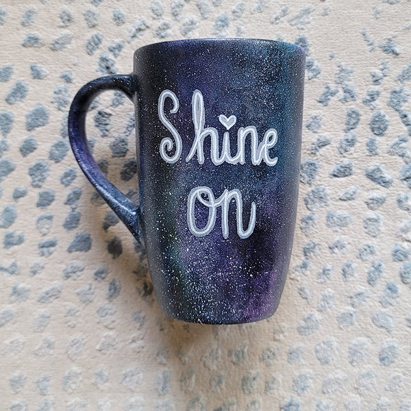 A Black galaxy hand painted mug with the words "Shine on"