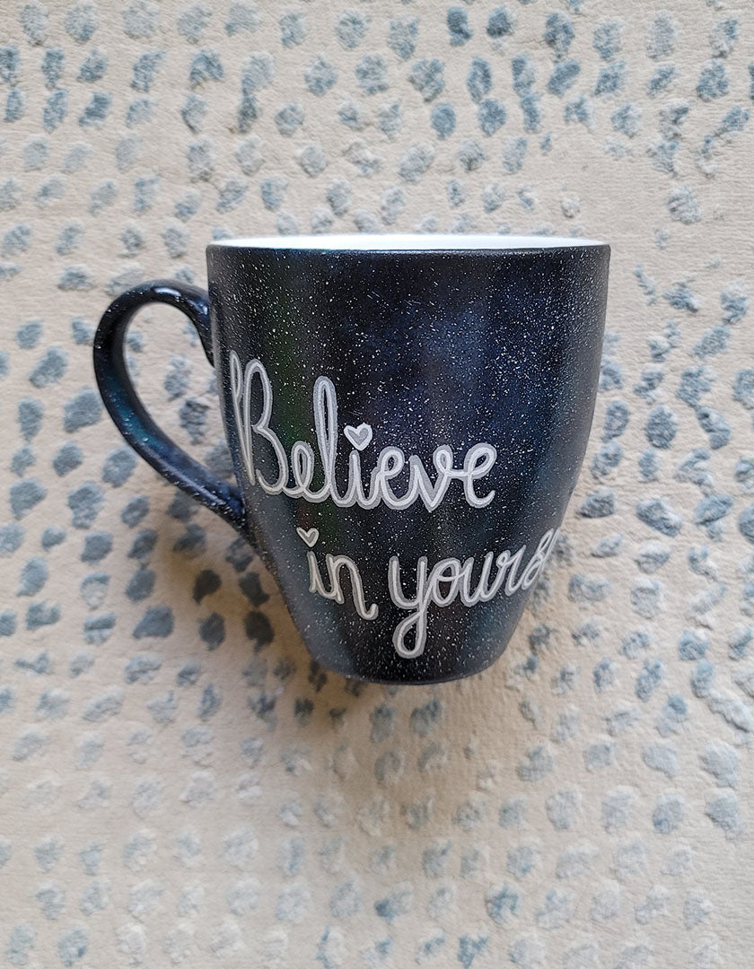 A Black galaxy hand painted mug with the words "Believe in yourself"