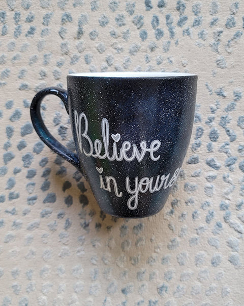 A Black galaxy hand painted mug with the words "Believe in yourself"