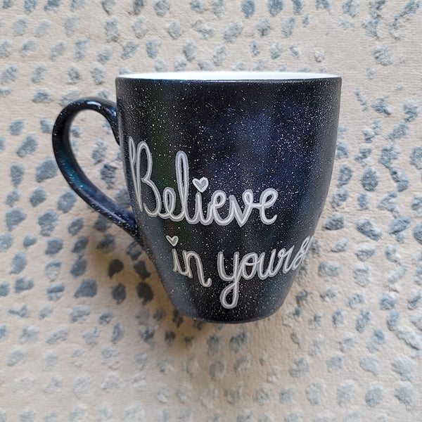 A Black galaxy hand painted mug with the words "Believe in yourself"