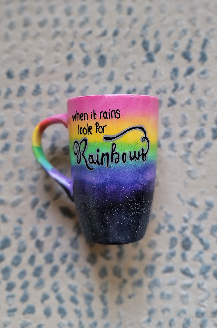 A Mug with the top half painted in rainbow and the bottom in dark galaxy, with the words on the rainbow written "when it rains look for rainbows"