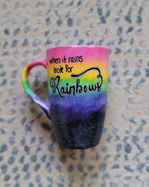 A Mug with the top half painted in rainbow and the bottom in dark galaxy, with the words on the rainbow written "when it rains look for rainbows"